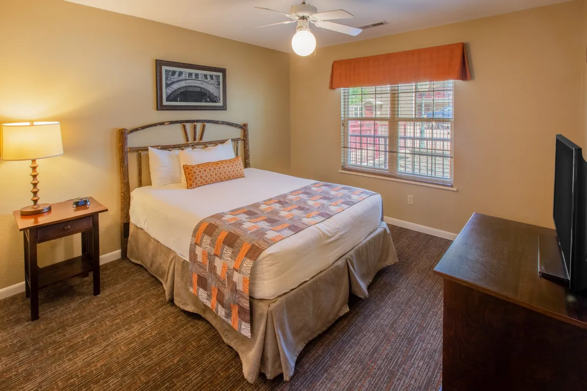 An inviting bedroom interior featuring a cozy bed, a nightstand with a lamp, a window allowing natural light to brighten the space, and a wall-mounted TV. The ambiance is restful and comfortable, suggesting a relaxing retreat after active days outside.