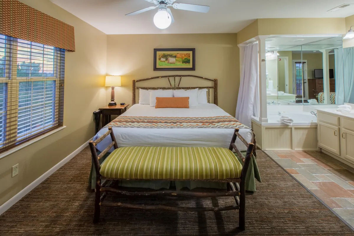 A serene bedroom featuring a large bed (likely a king), bench, bedside lamp, and framed artwork. The adjoining bathroom area is partially visible, emphasizing comfort and relaxation, with natural light streaming in.
