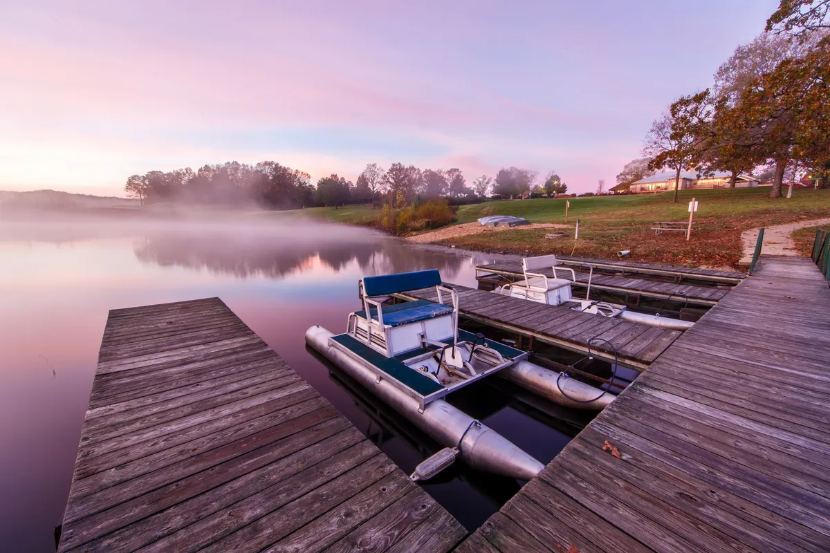 A scenic view of a dock stretching out onto the lake, with boats moored nearby and trees framing the scene. The tranquil water and inviting dock present opportunities for boating, fishing, and enjoying quiet moments by the lake.
