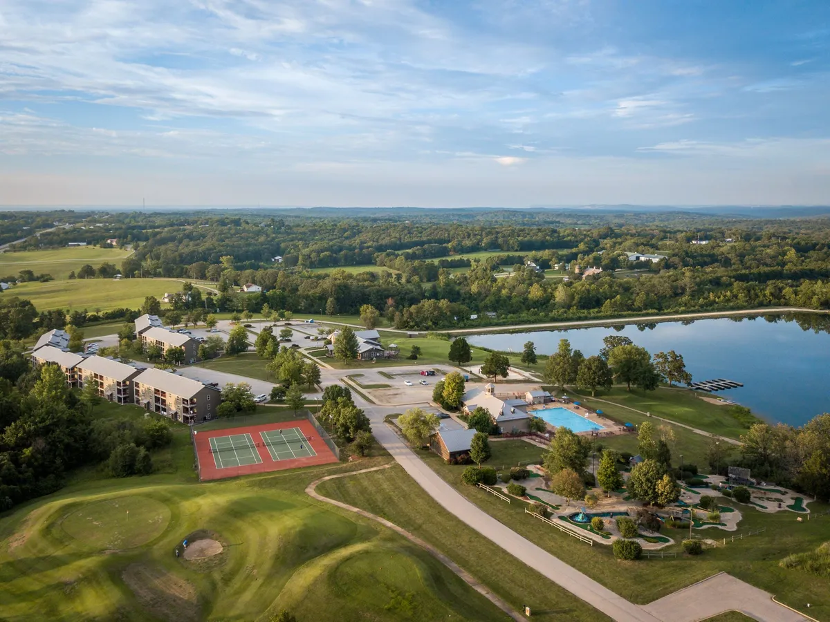 A panoramic view of the resort’s extensive outdoor amenities, including tennis courts, a large swimming pool, lakeside areas, and residential buildings nestled in greenery. The scene is vibrant and active, suggesting a range of recreational options just steps away.