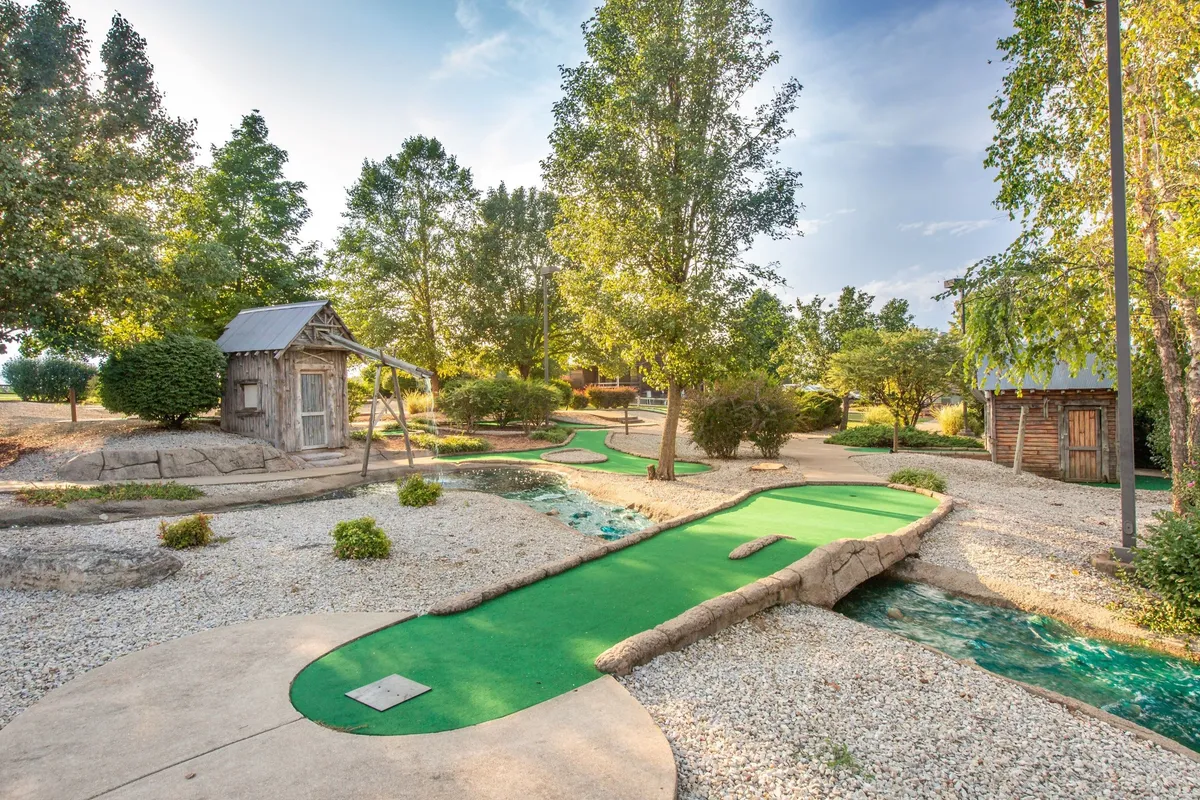 A bright and playful outdoor mini-golf course set amid lush greenery, with a swing set and wooden play structures nearby. Families and children can be imagined enjoying a cheerful day outdoors, competing in mini-golf or playing on the swings under the sun.