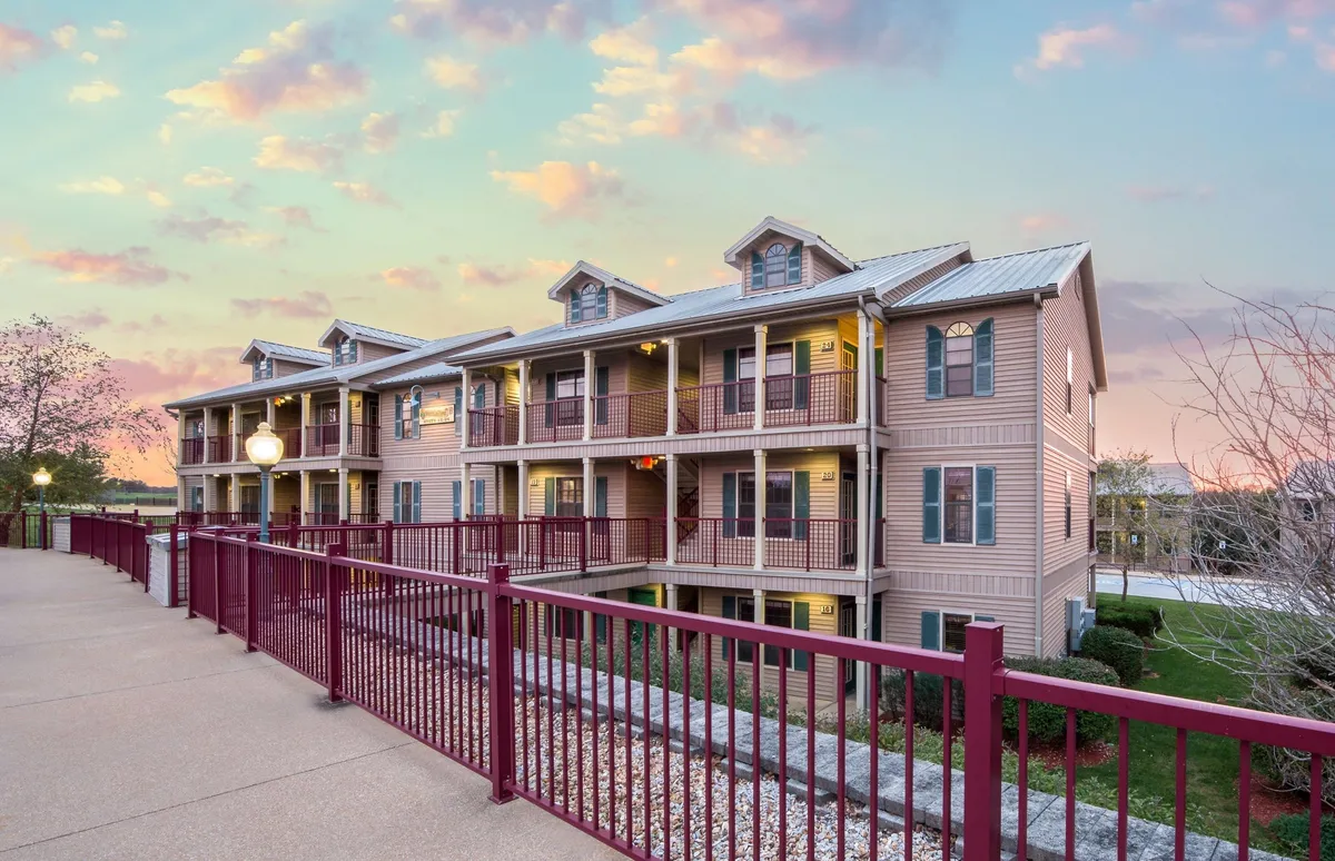 This exterior image showcases the facade of the resort with inviting balcony railings overlooking a landscaped communal area. Warm lighting fixtures create a welcoming atmosphere, suggesting evening activities and relaxation. The scene hints at outdoor enjoyment and social gatherings, set against the backdrop of a beautiful outdoor environment.